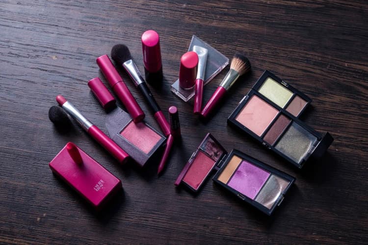 Um conjunto de produtos de maquiagem, incluindo batons e paletas de sombras, dispostos artisticamente sobre um fundo texturizado de madeira escura.
