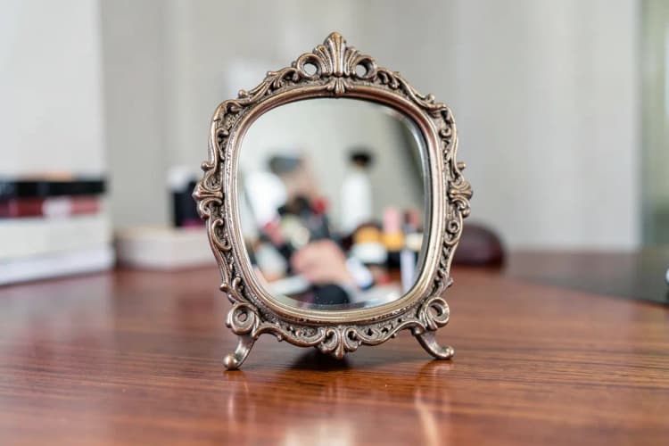 Um espelho de maquiagem vintage com uma moldura ornamentada, refletindo um leve borrão de um ambiente de beleza, sobre uma mesa de madeira.