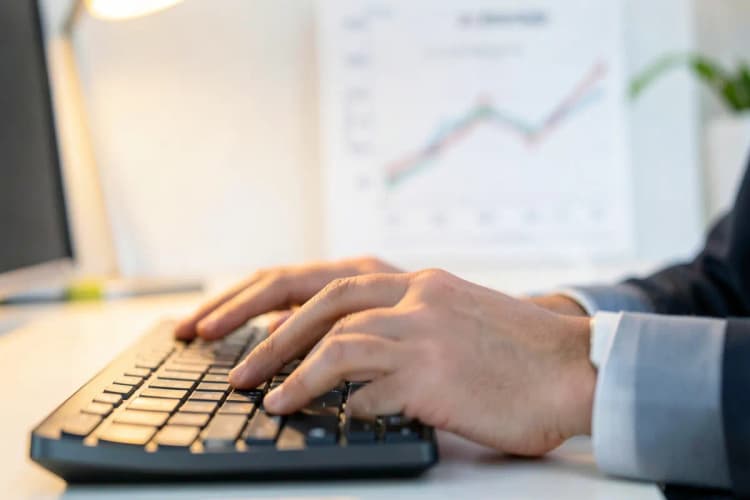 Mãos digitando em um teclado de computador com um gráfico de crescimento ao fundo.